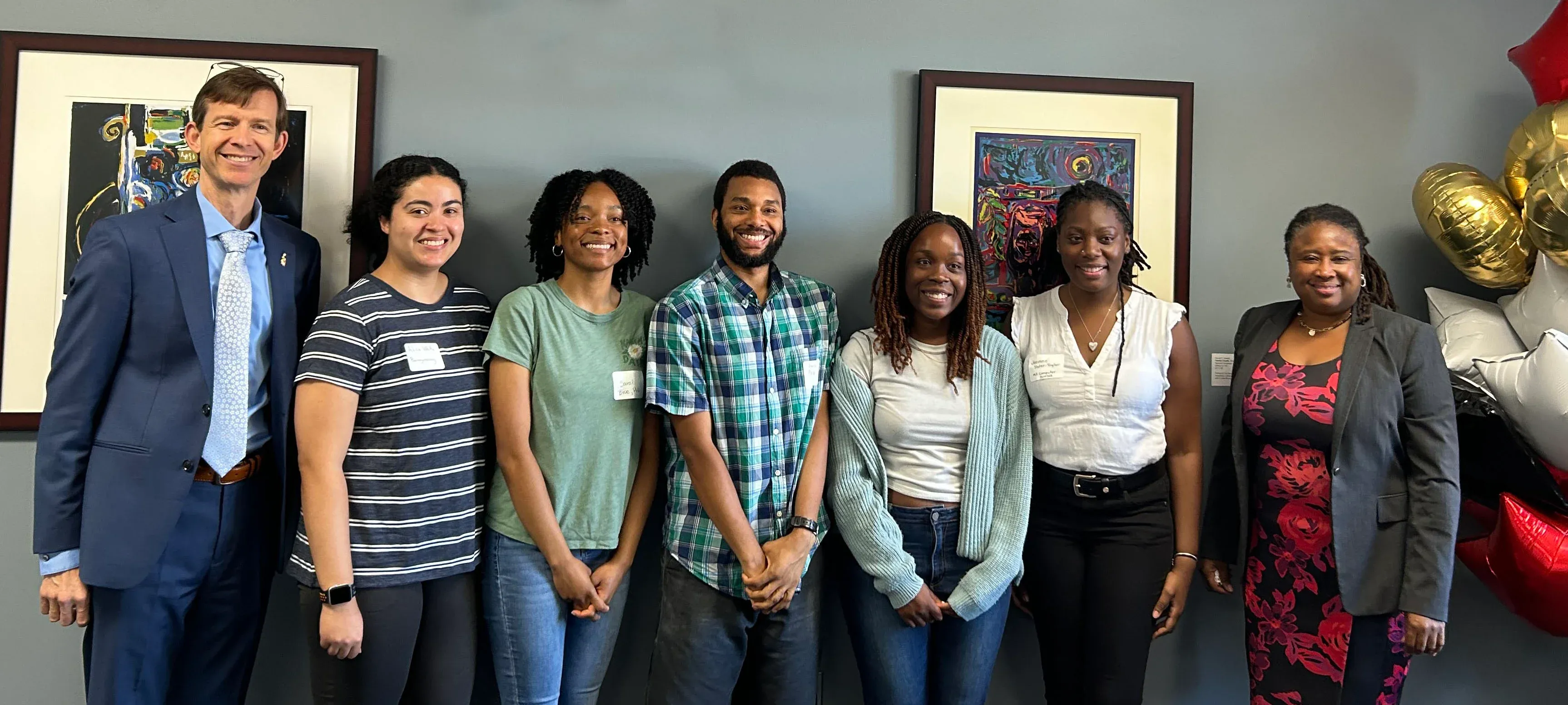Dean Roth and Dr. Evelyn Cooper standing with a group of GEM Fellows in front of a gray wall with balloons nearby.